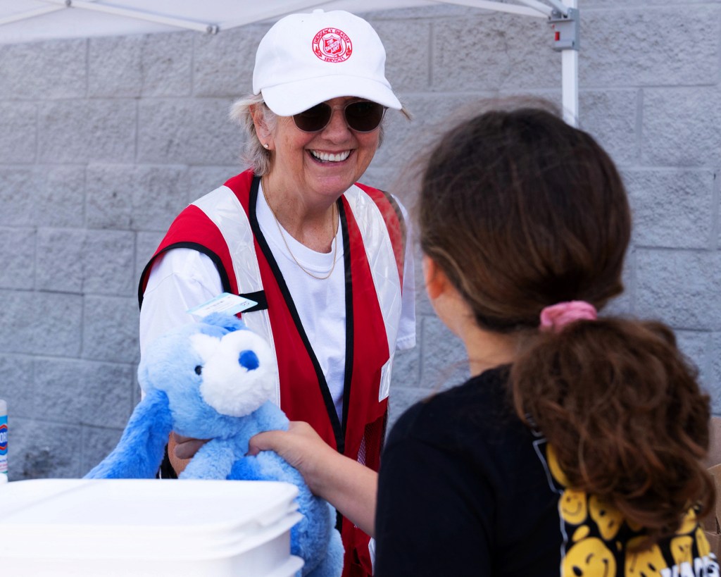 A smiling volunteer wearing a red vest and a baseball cap hands a blue stuffed animal to a young girl, who is reaching out to receive it.