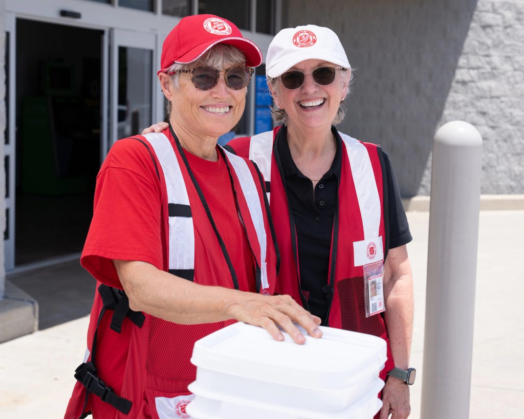 Two female volunteers from The Salvation Army smile while holding containers outside a Walmart during a disaster response operation.