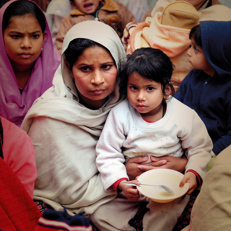 A concerned mother holds her child in her lap, both looking somber. The mother is wearing a light-colored headscarf and an outer garment, while the young girl in her arms is dressed in a white sweater, holding a bowl and spoon.