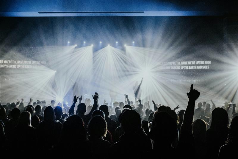 A large crowd of people at a concert, silhouetted against bright stage lights, with some audience members raising their hands in excitement. Text on the screen in the background suggests a worship or spiritual theme.