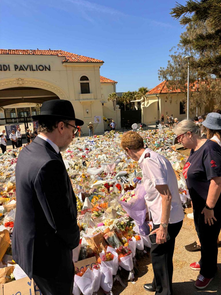 A group of people examining a large memorial made of flowers and tributes outside Bondi Pavilion, with the building in the background.