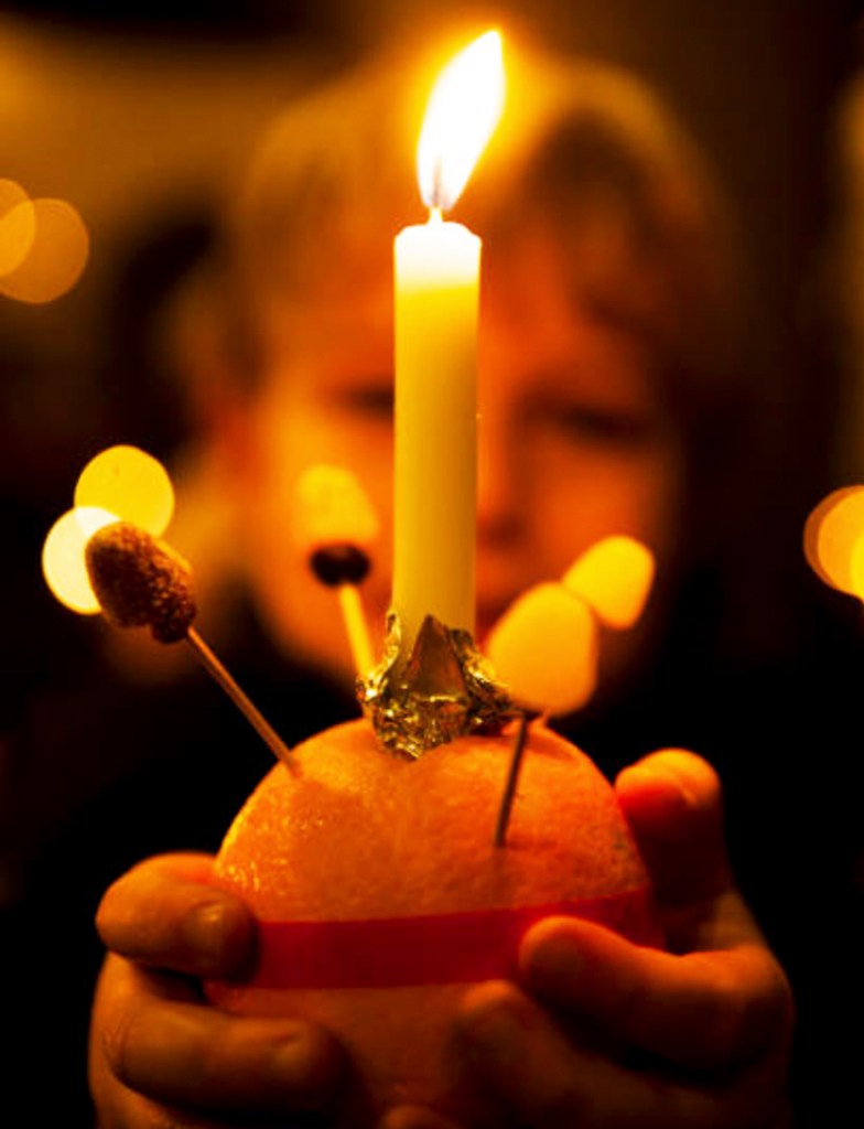A child holding a Christingle orange with a lit candle on top, surrounded by festive decorations and warm lights.