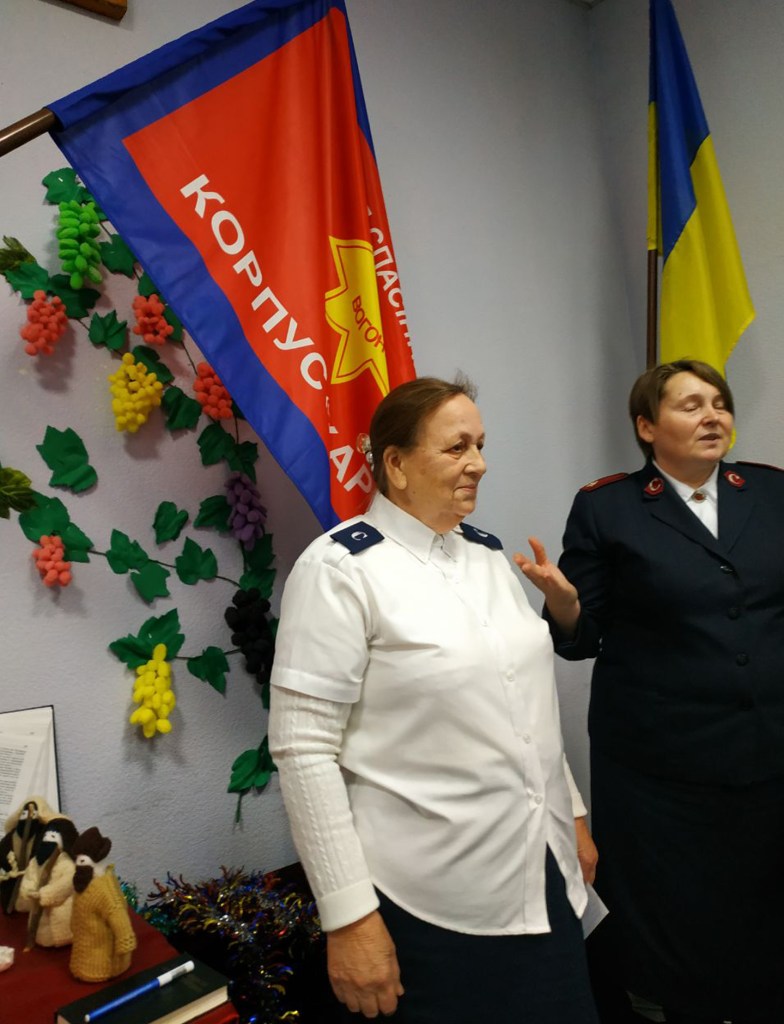 Two women in Salvation Army uniforms stand in front of a colorful banner, while a decorated wall displays vibrant artificial grapes. The setting conveys a sense of community and support.