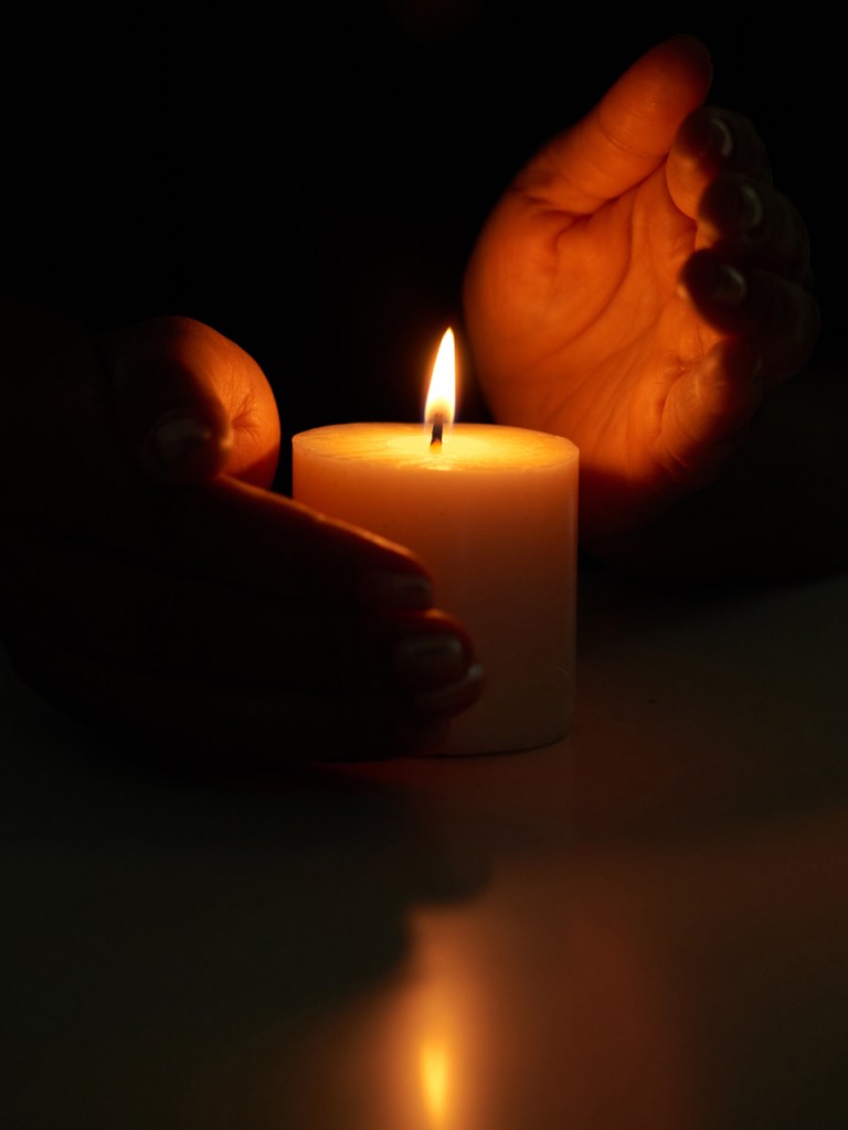 A lit candle held gently between two hands in low light, creating a warm, calming atmosphere.
