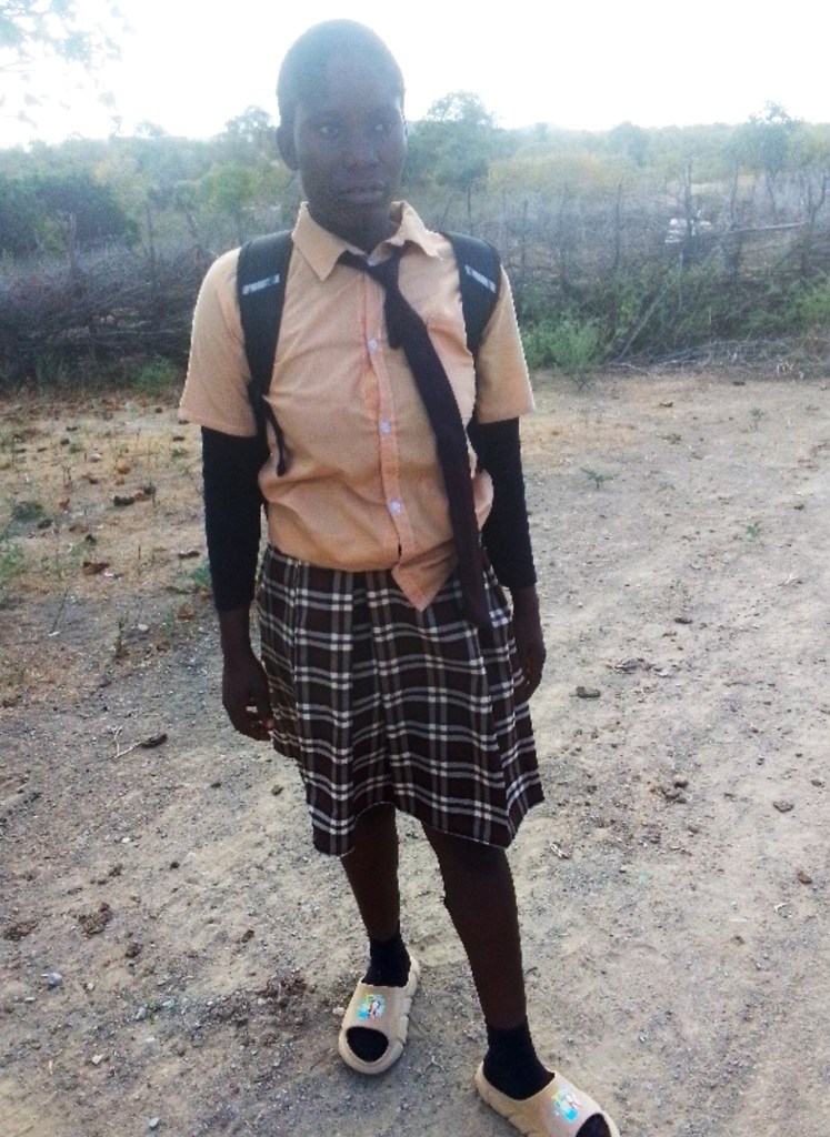A student standing on a dirt path, wearing a short-sleeved, light orange shirt, a tie, and a brown checkered skirt. The student has a black backpack, black socks, and light-coloured sandals. The background features sparse vegetation and a barbed-wire fence.