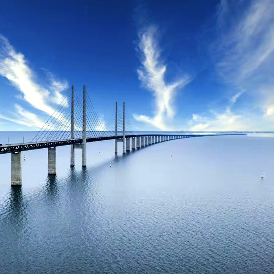 A panoramic view of a cable-stayed bridge stretching across calm water under a clear blue sky with wispy clouds.