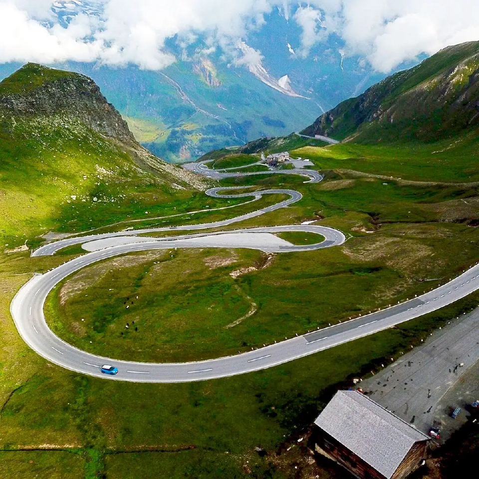 Aerial view of a winding mountain road surrounded by green hills and snow-capped peaks under a cloudy sky.