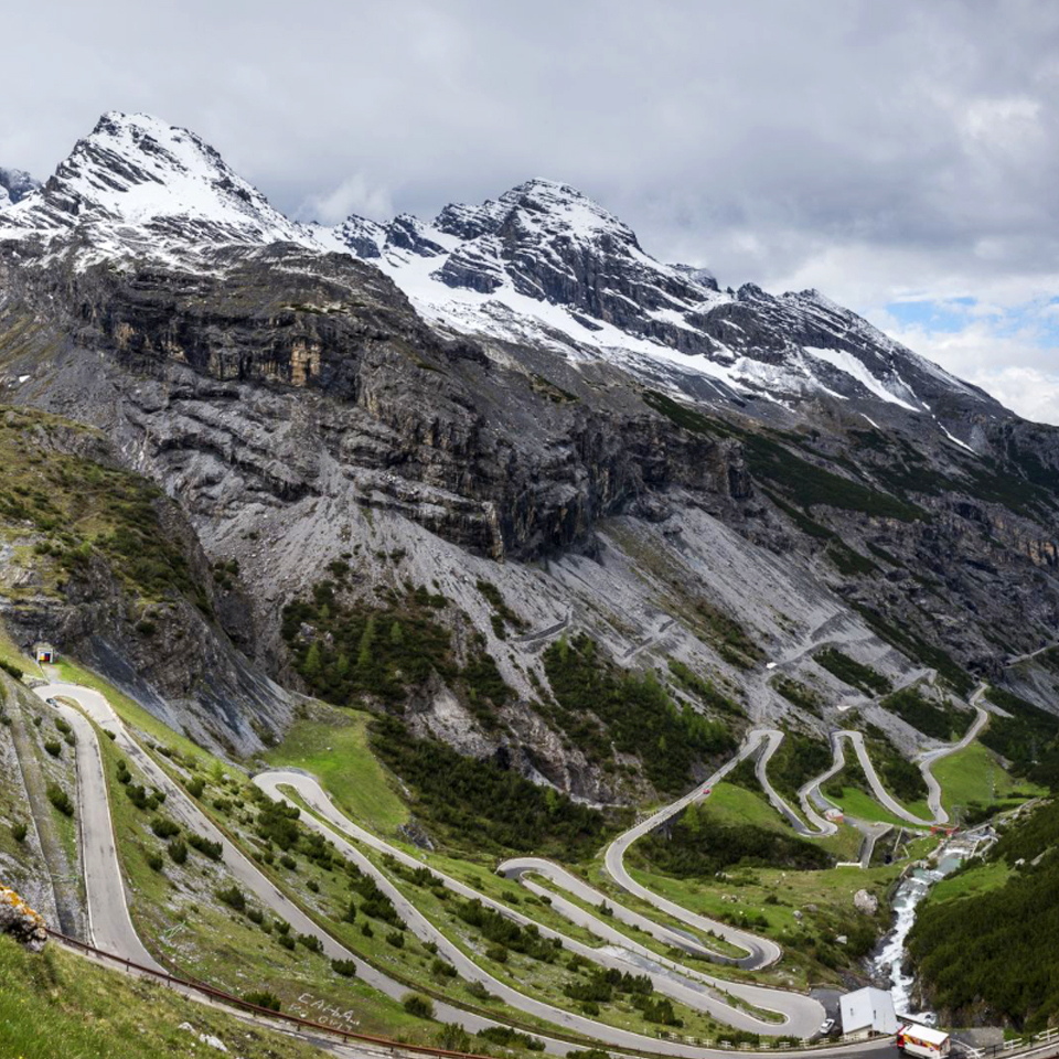 A panoramic view of a winding mountain road with lush green landscapes and rocky cliffs, surrounded by snow-capped peaks under a cloudy sky.