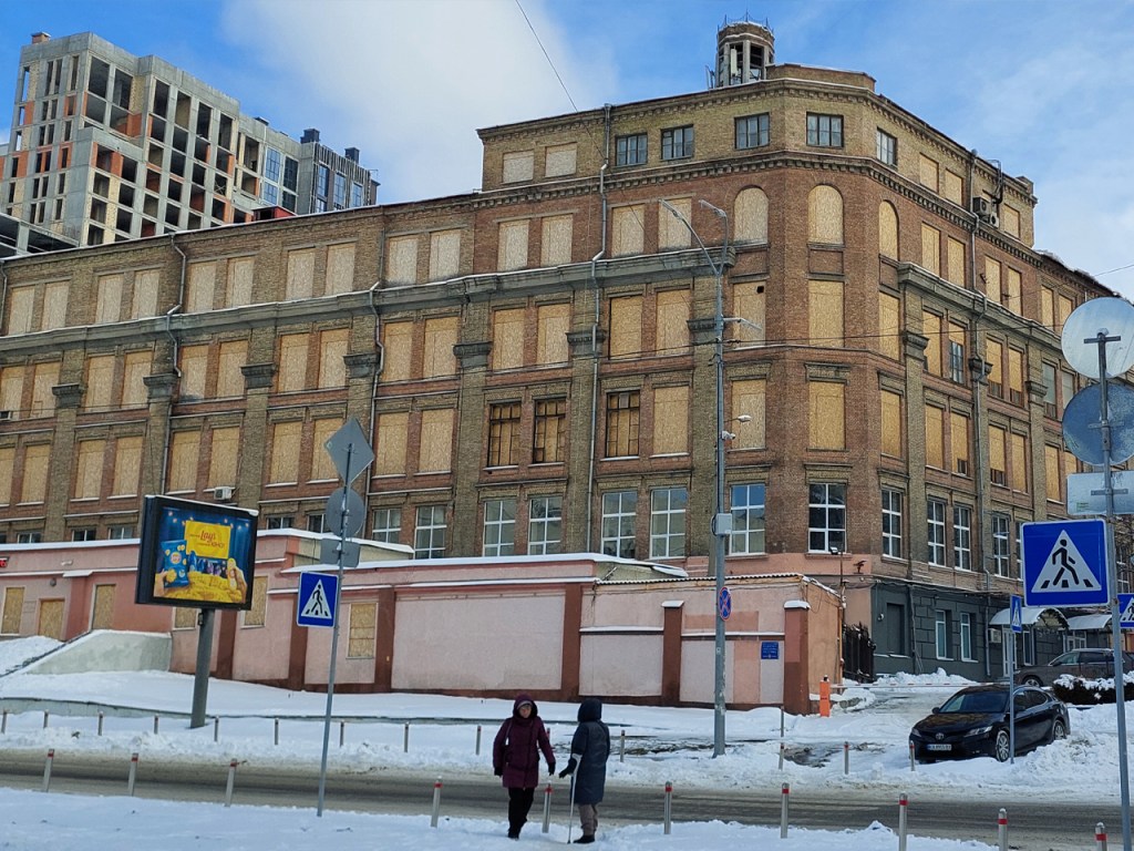 A large, abandoned brick building with boarded-up windows, situated near a snowy street. In the foreground, two figures walk past traffic signs and a billboard, while a modern construction site is visible in the background.