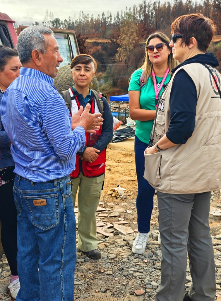A group of five individuals engaged in conversation outdoors, with a landscape featuring burnt trees in the background. The man in a blue striped shirt is speaking, while the others, including women in varying attire, listen intently.