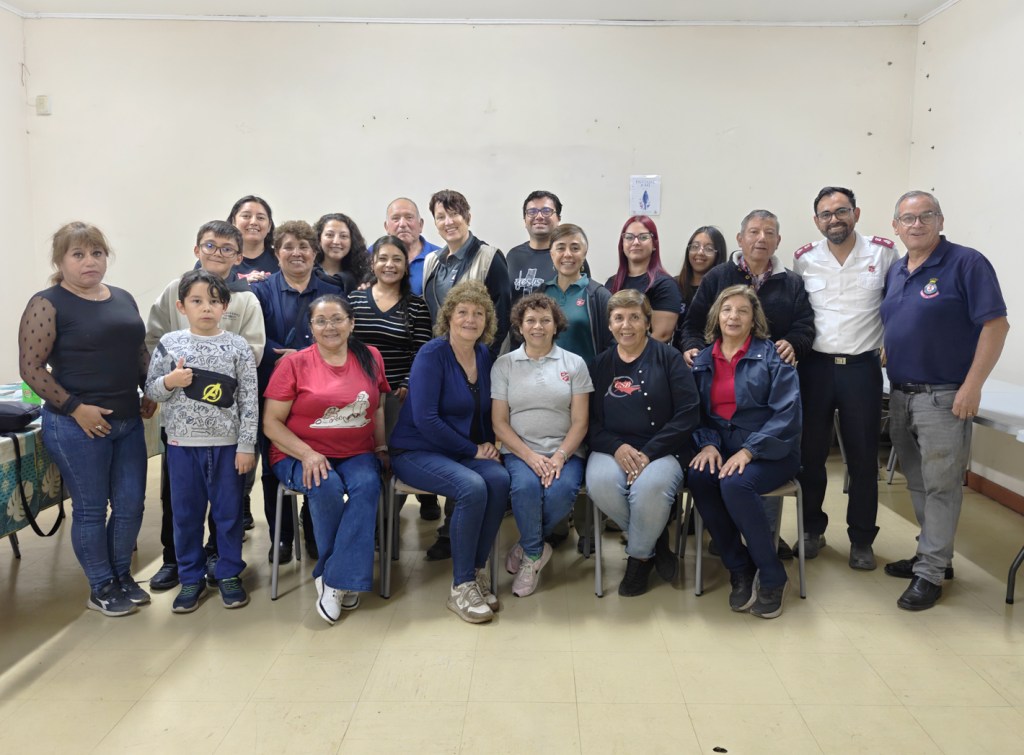 A diverse group of people, including men, women, and children, posing together in a community hall setting. They are smiling and standing or sitting in rows, wearing casual clothing. The atmosphere is friendly and welcoming.