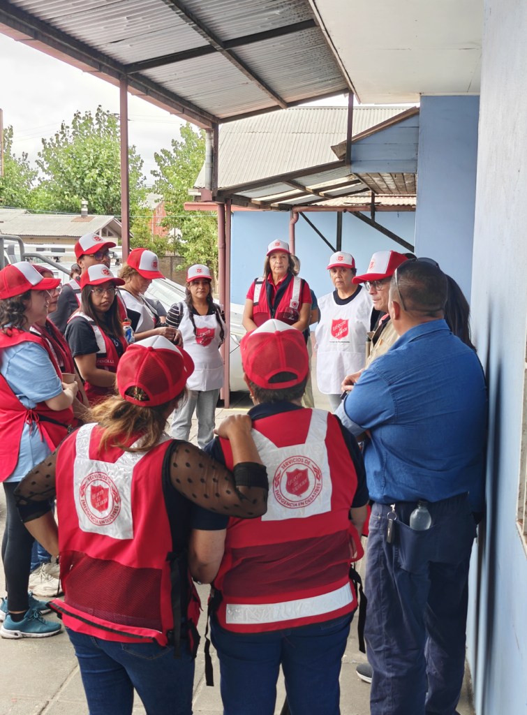 A group of volunteers wearing red vests and caps, gathered outside under a covered area, listening to a speaker.