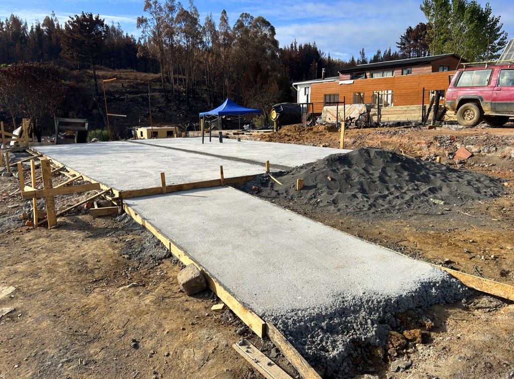 A concrete foundation slab is under construction, surrounded by wooden frames with a pile of gravel nearby. In the background, there are partially burnt trees and a residential building.