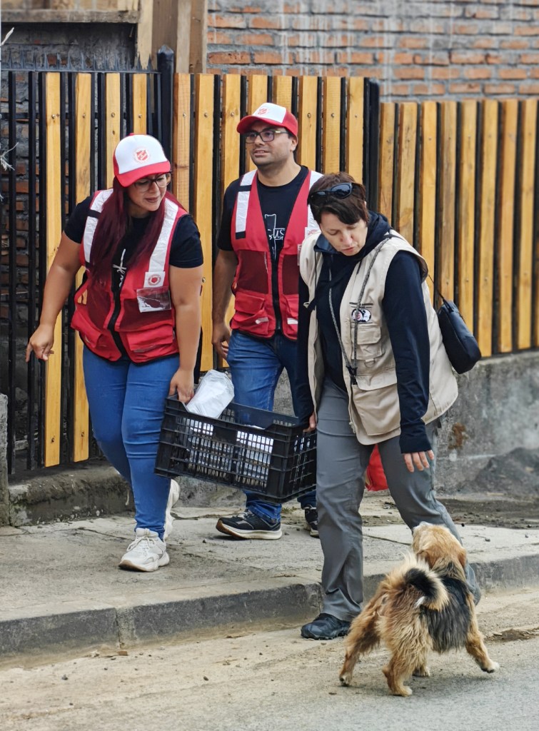 Three individuals in red vests walk along a street, one carrying a basket. A small dog approaches them.