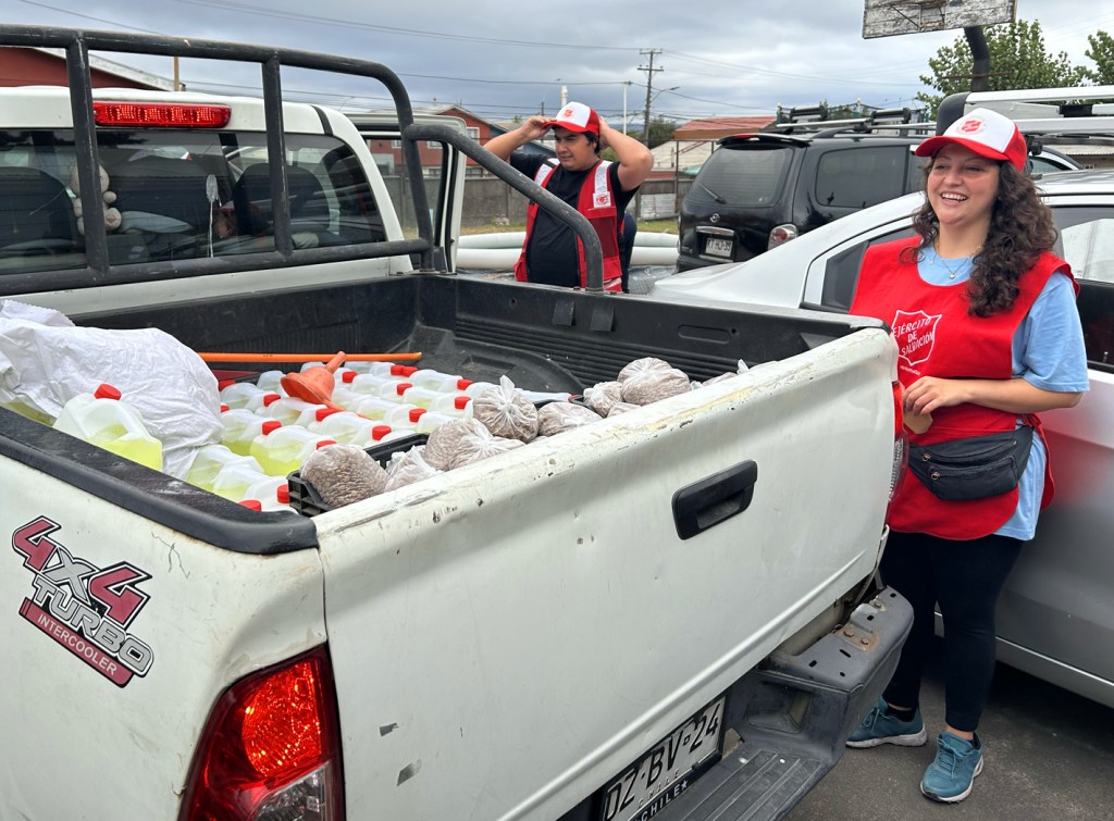 Two volunteers in red vests preparing supplies in the back of a pickup truck, with bottles and bags visible, set against a cloudy sky.
