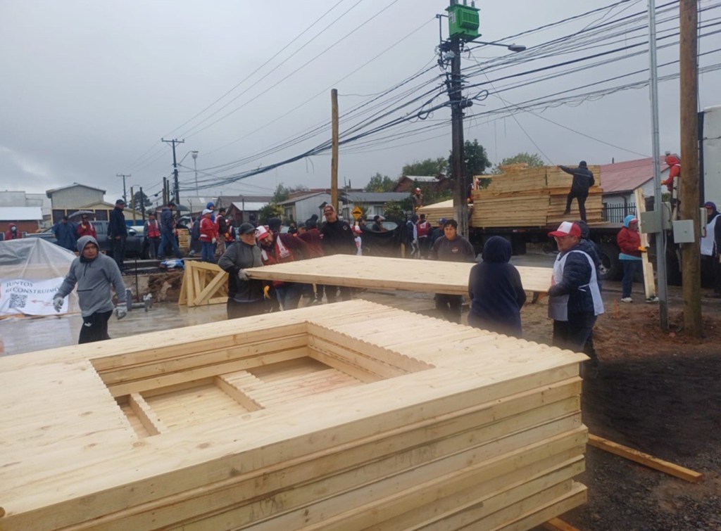 Workers collaborating to assemble wooden panels at a construction site, with stacks of lumber and power lines in the background on a cloudy day.