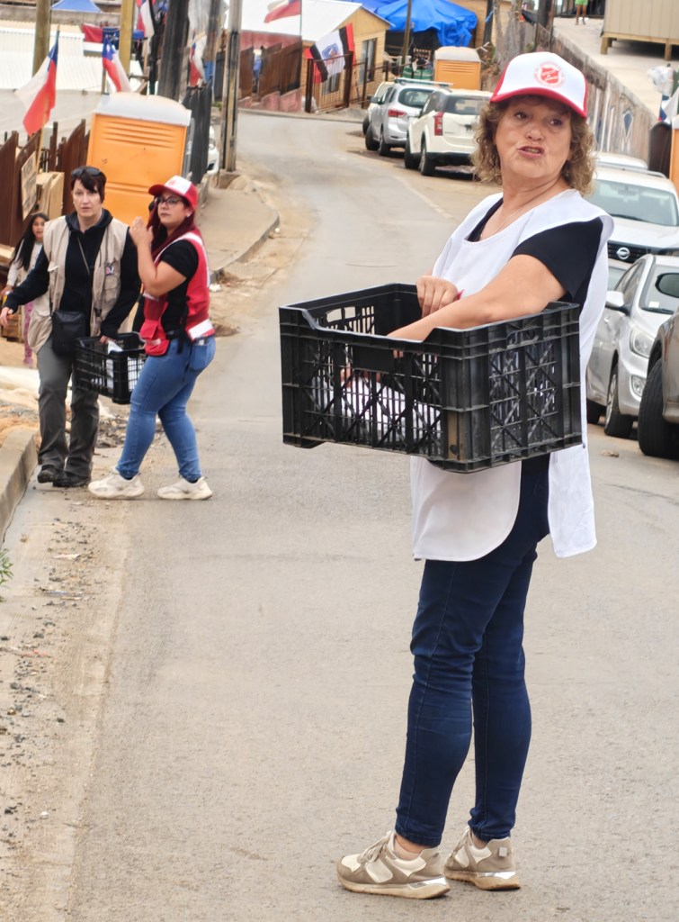 A woman holding a crate while wearing a white vest and a red cap, with two other individuals in the background, on a street decorated with flags.