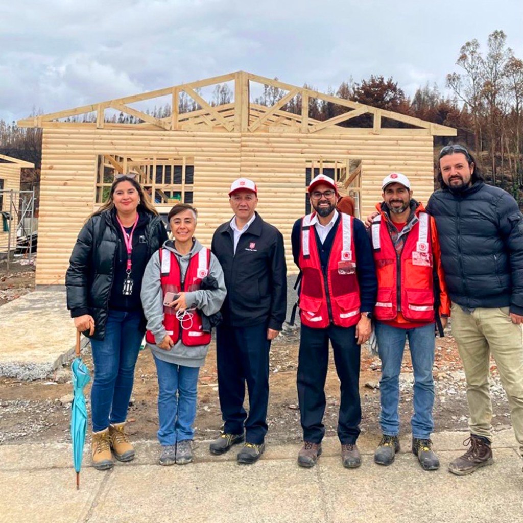 A group of six individuals standing in front of a partially constructed wooden house, with a cloudy sky in the background. The people are wearing red vests, with some holding umbrellas, and are smiling at the camera.