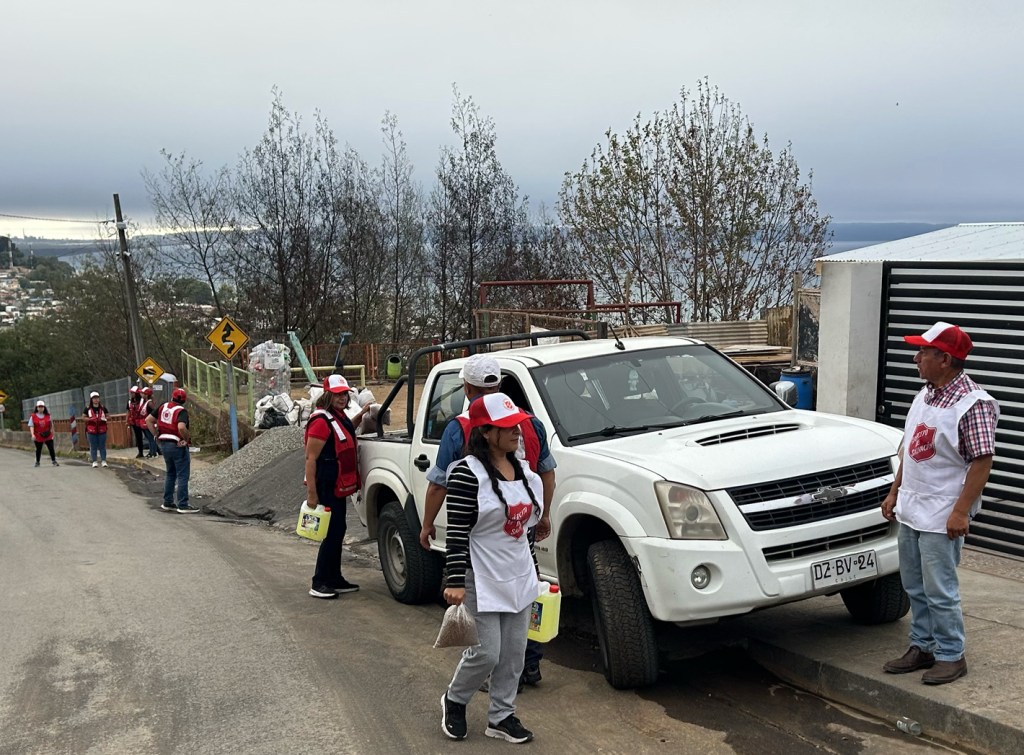 A group of individuals in red vests and white aprons stand on a road beside a parked white vehicle, with some people holding containers. The background features trees and a winding street sign, indicating a winding road.