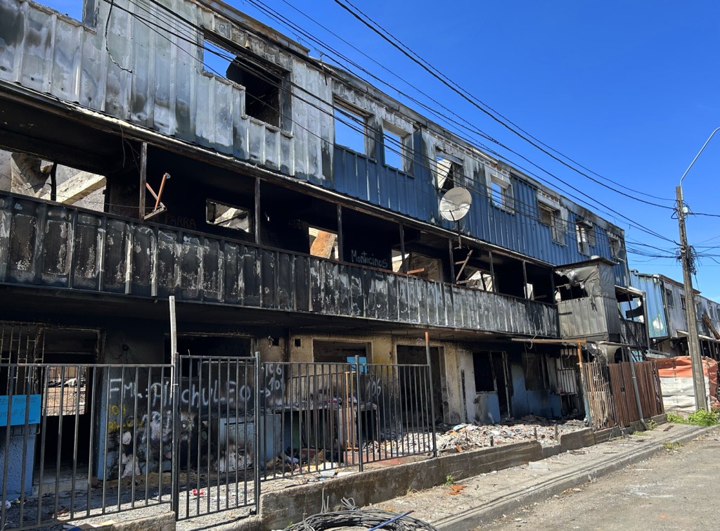 A charred and abandoned building with damaged windows and a burnt exterior, surrounded by debris and a metal fence, under a clear blue sky.