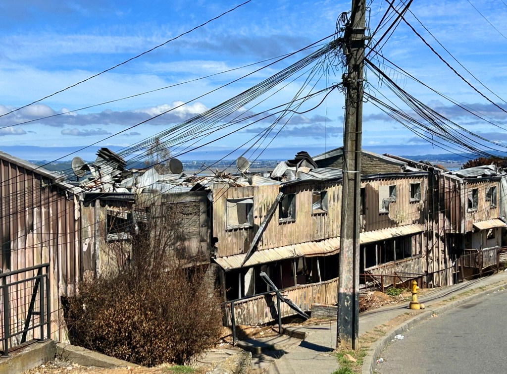 A view of a rundown area featuring dilapidated buildings with peeling paint and damaged roofs, surrounded by tangled power lines and a clear blue sky.