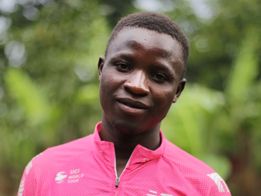 A young man wearing a pink cycling jersey with 'UCI World Tour' printed on it, standing outdoors with greenery in the background.
