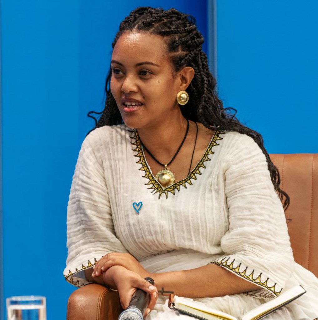 A woman with braided hair wearing a traditional white dress with gold detailing, sitting on a brown chair. She is holding a microphone and appears to be engaged in conversation.