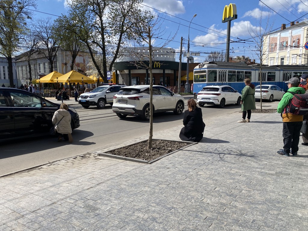 A busy street scene with cars and a tram, featuring people on the pavement including two individuals sitting. In the background, a McDonald's restaurant with yellow umbrellas and a blue sky.