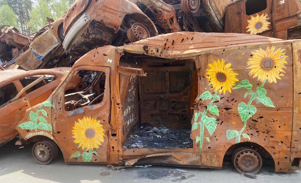 A rusted, abandoned van partially hidden among other derelict vehicles, featuring vibrant sunflower artwork on the sides.