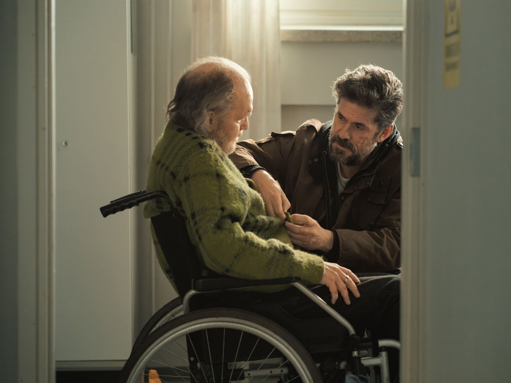 A caregiver gently assists an elderly person in a wheelchair, helping with their clothing in a softly lit room.