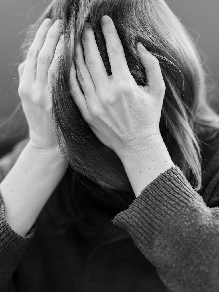 A person with long hair, shown from the shoulders up, is holding their head in their hands, conveying a sense of distress or anxiety. The image is in black and white.