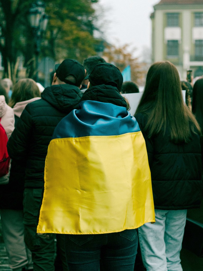 A person wearing a blue and yellow flag wrapped around their shoulders, standing among a crowd in an outdoor setting.