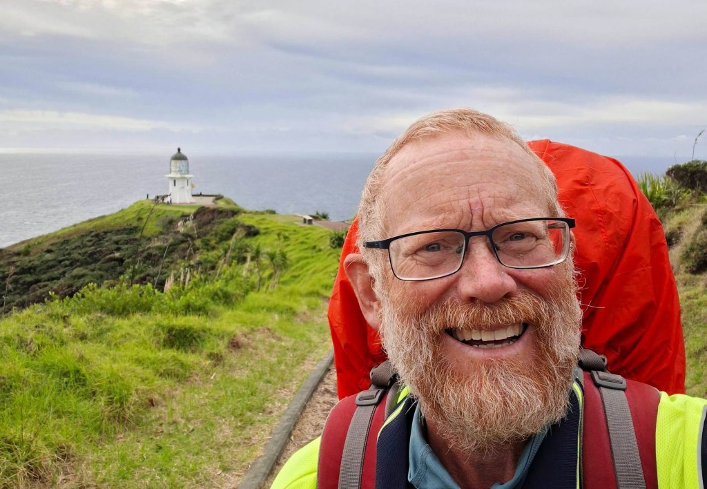 A smiling elderly man with a beard and glasses stands in front of a lighthouse on a hill, with the ocean in the background, while carrying a red backpack.