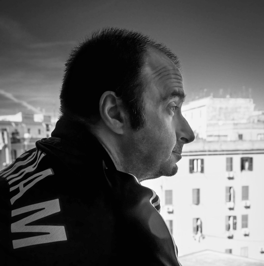 A contemplative man in profile, gazing out of a window, with buildings visible in the background. The image is in black and white.