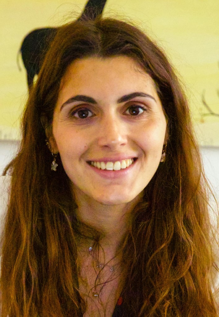 Close-up portrait of a woman with long, wavy brown hair and dark eyes, smiling warmly at the camera, set against a softly painted background.