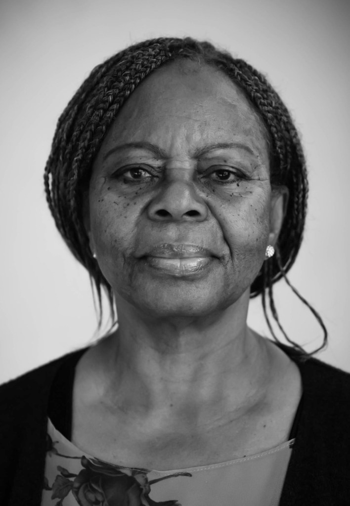 Black and white portrait of an elderly woman with braided hair, wearing a floral top and a black cardigan, looking directly at the camera with a neutral expression.