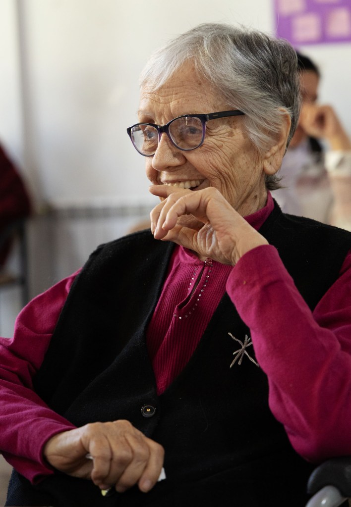 An elderly woman with grey hair and glasses smiling while resting her chin on her hand, wearing a pink top and a black vest.