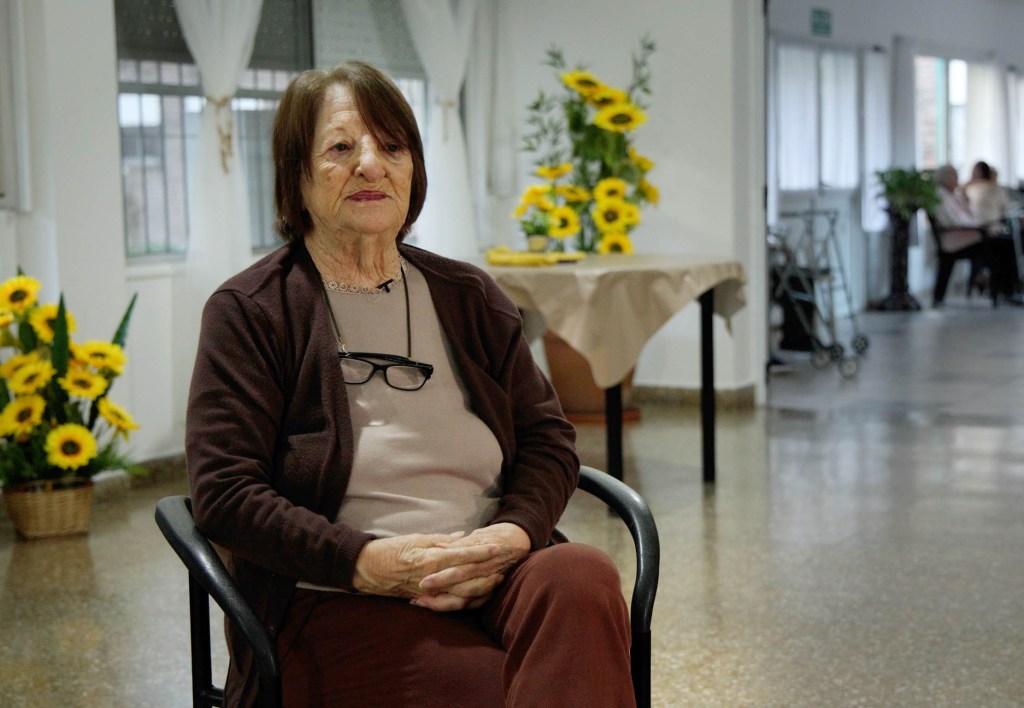 An elderly woman sitting in a chair, wearing a brown cardigan and glasses, in a bright room decorated with sunflowers.