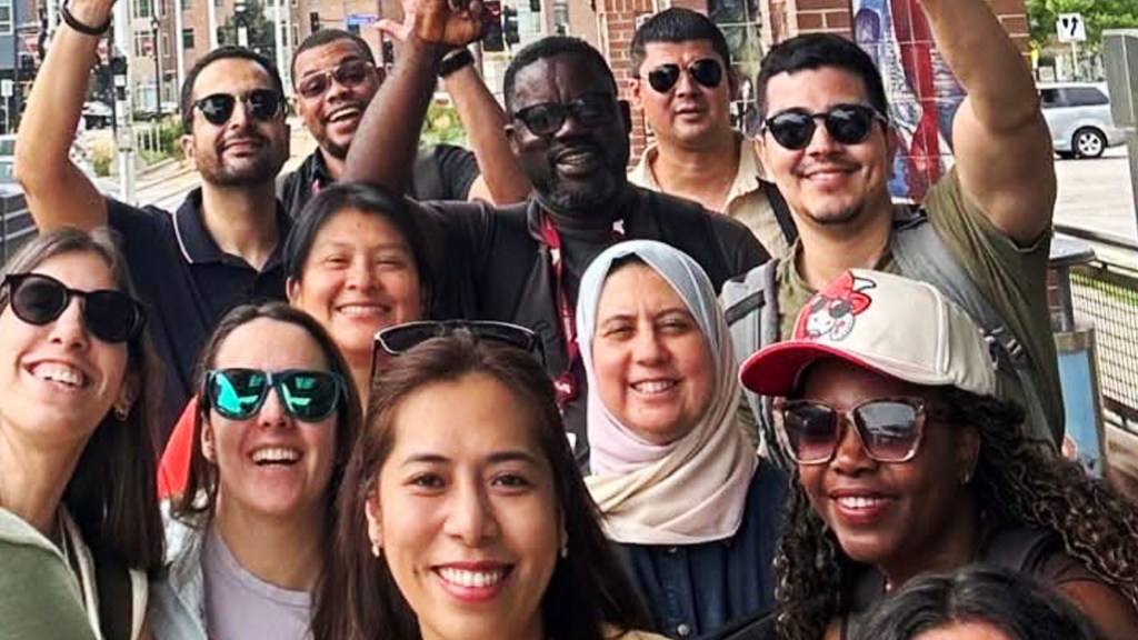 A diverse group of people smiling and posing for a photo outdoors, with some wearing sunglasses and casual clothing. They seem to be enjoying their time together, with a few individuals giving a thumbs-up and peace signs.