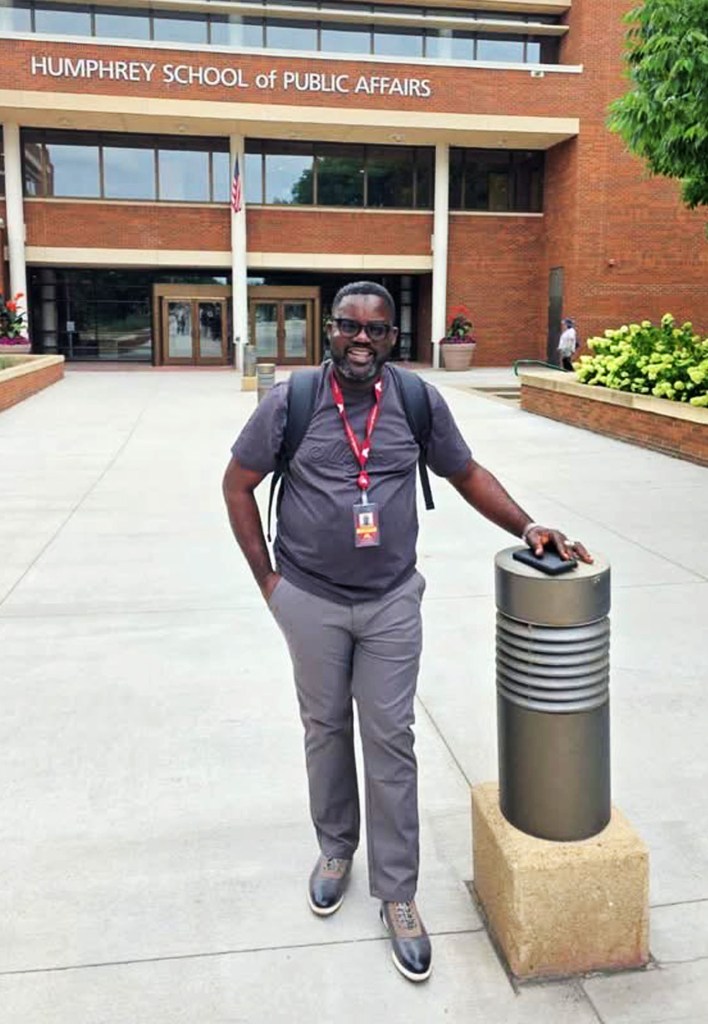 A smiling man stands in front of the Humphrey School of Public Affairs, wearing glasses, a grey shirt, and grey trousers, with a backpack and a lanyard around his neck.