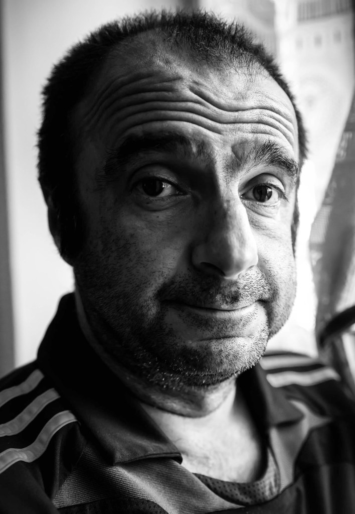 A close-up portrait of a middle-aged man with short hair and a light beard, wearing a dark sports jersey. The image is in black and white, highlighting his facial features and expression, against a softly blurred background.