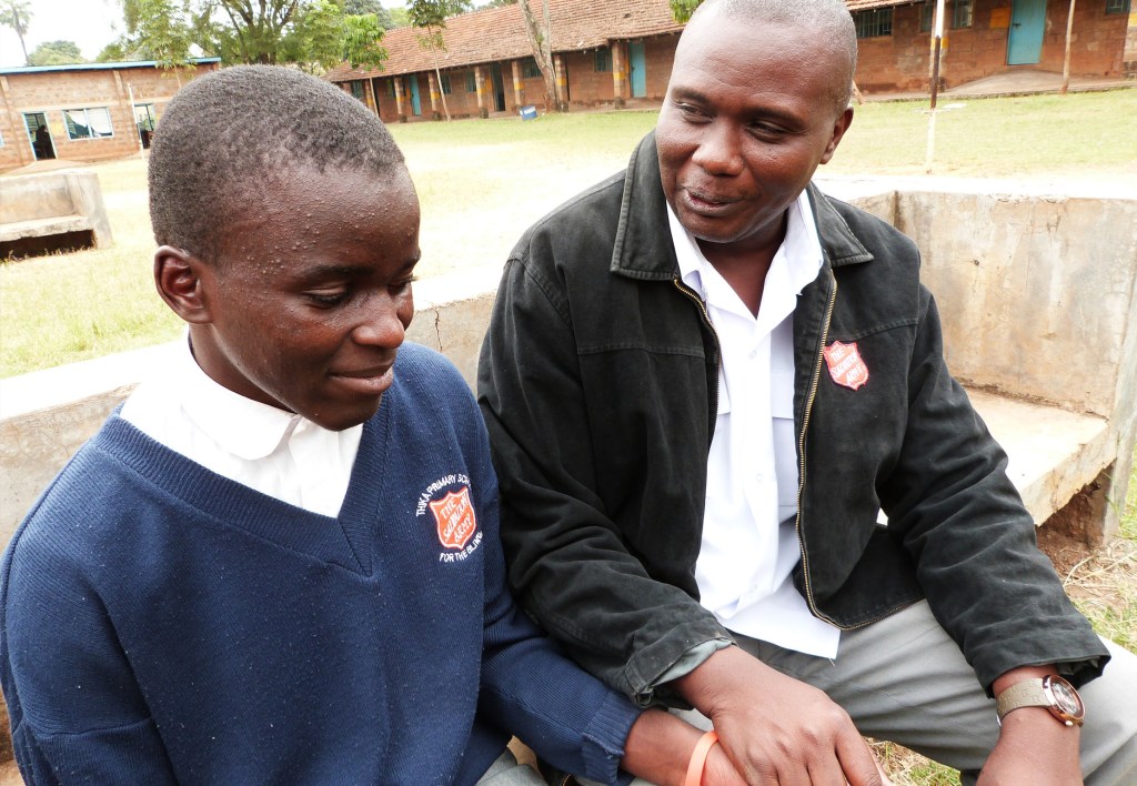 A young student in a blue sweater sits next to an adult male mentor, both engaged in a conversation outdoors. The background features a school setting with buildings and green grass.